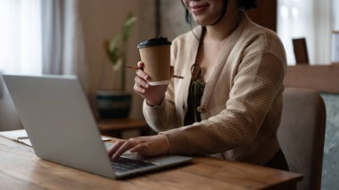 Güzel ve mutlu bir Asyalı kadının, bir kafede uzaktan çalışan, bir masada dizüstü bilgisayarıyla çalışırken kahvesinin tadını çıkaran görüntüsü. Kentsel yaşam tarzı konsepti