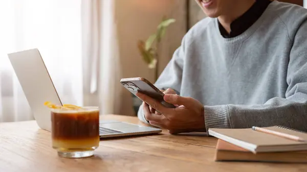 A happy Asian man in a sweater enjoys chatting with his friends on his phone, using his smartphone while working remotely at a coffee shop. Lifestyle concept