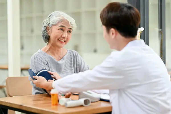 A happy Asian retired old lady is being measured for her heart rate and blood pressure with a monitor while talking with a doctor in an examination room in a hospital.