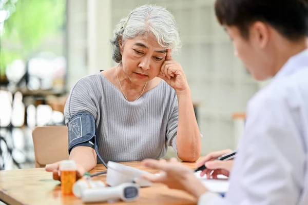 A stressed and concerned Asian retired old lady is being measured for her heart rate and blood pressure with a monitor while talking with a doctor in an examination room in a hospital.