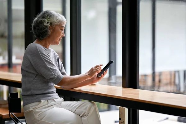 A happy Asian retired lady in casual clothes is using her smartphone at a table while relaxing in a coffee shop in the afternoon. Lifestyle and technology concepts