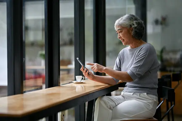 A relaxed and happy Asian retired lady in casual clothes enjoys the app on her smartphone while sitting in a coffee shop. Retirement lifestyle concept