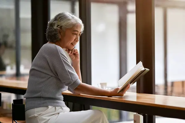 A happy and relaxed Asian retired lady is reading a book while sitting at a table in a coffee shop, spending her weekend in the city alone. Retirement lifestyle concept