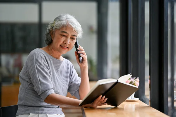A happy and relaxed Asian retired lady is reading a book and talking on her phone with someone while sitting in a coffee shop. Lifestyle concept