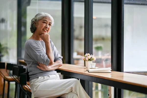 A portrait of a beautiful and happy Asian retired woman in casual clothes sits at a table by the window in a coffee shop. Happy retirement lifestyle concept