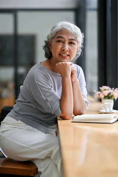 A portrait of a beautiful and happy Asian retired woman in casual clothes sits at a table by the window in a coffee shop. Happy retirement lifestyle concept