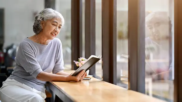 A happy and relaxed Asian retired lady is enjoying reading a book while sitting in a coffee shop. Lifestyle concept