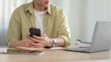 Sıradan giyinmiş yakışıklı bir Asyalı 'nın görüntüsü bilgisayarındaki bir şeye bakarken akıllı telefonunu tutuyor..