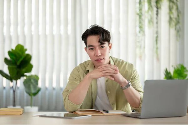 A portrait of a handsome and attractive young Asian man in casual clothes looks at the camera and sits at a table with his laptop, tablet, and books on the table.