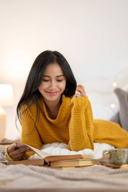 Mutlu ve çekici Asyalı bir kadının portresi rahat örülmüş bir süveter içinde abur cubur yemekten ve bir sonbahar günü yatağında kitap okumaktan zevk alır. Boş zaman konsepti