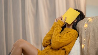 Yatak odasında kitap okurken uykulu ya da yorgun hisseden, rahat örülü süveter giyen, yüzünü kitapla örten Asyalı bir kadının yan görüntüsü..