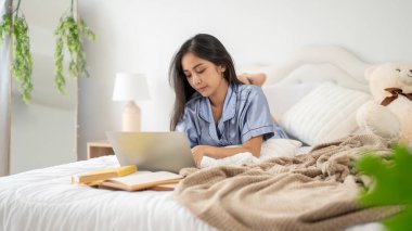 Pijamalı güzel bir Asyalı kadın yatak odasında yatağında yatarken laptopuyla işine odaklanıyor..