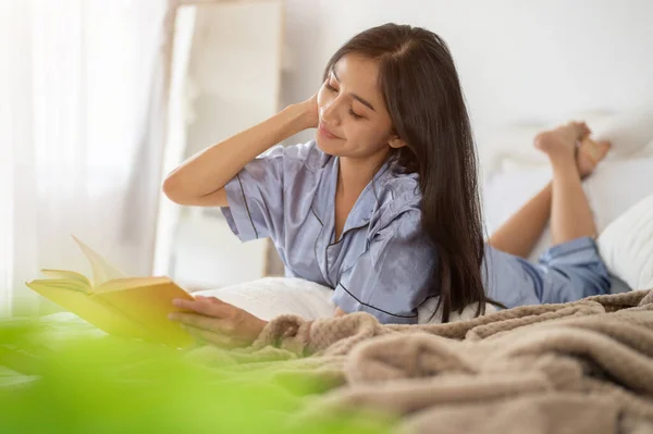 stock image An attractive and happy Asian woman in pajamas relaxes reading a book on her bed in the bedroom in the morning. Hobby and leisure concept