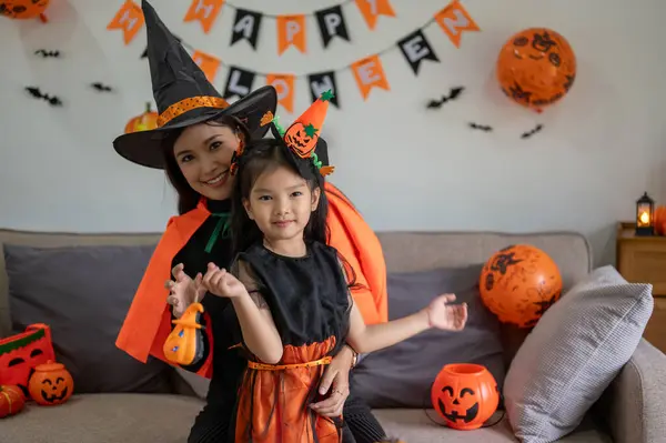 A beautiful Asian mom is embracing her daughter while celebrating Halloween in the living room together. Mom and young daughter girl in Halloween costume