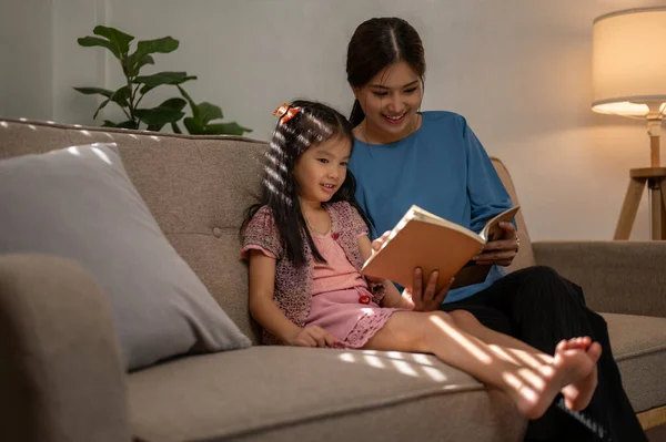 행복한 아시아의 엄마는 거실에서 소파에서 휴식을하면서 그녀의 딸에게 재미있는 읽기를 — 스톡 사진