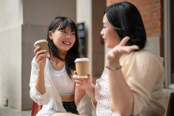 A beautiful young Asian woman enjoys talking and having coffee with her friend while sitting on the stairs in the city. Urban lifestyle concept