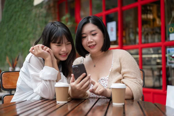 Two happy young Asian female friends are watching a video on a smartphone together while relaxing at an outdoor table of a coffee shop in the city.