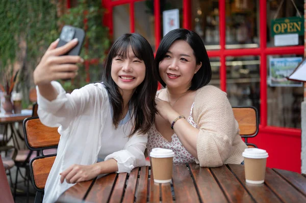 Two happy young Asian female friends are using a smartphone to take selfies together while relaxing at an outdoor table of a coffee shop in the city.