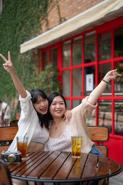 Two cheerful and happy young Asian female friends are sitting at an outdoor table in a coffee shop together, raising their hands, and enjoying the weekend together. Lifestyle and friendship concepts