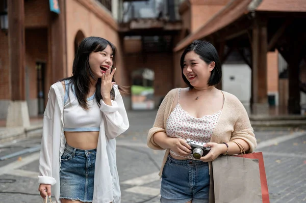 Two cheerful and happy young Asian female friends are enjoying talking and having fun while walking around the city on a shopping day on the weekend together. Lifestyle concept