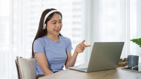 A cheerful young Asian woman in headphones is talking on an online meeting or online job interview while sitting at a table at home.