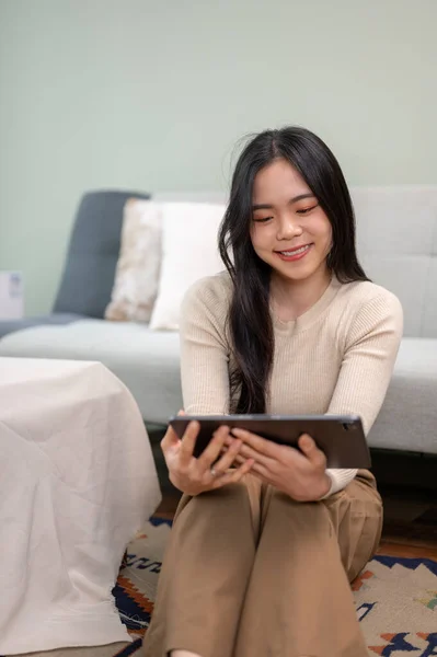 A beautiful young Asian woman watching a movie or video on her digital tablet while relaxing in her living room. People and leisure concepts