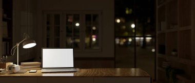 Beyaz ekran dizüstü bilgisayar, masa lambası ve gece modern karanlık bir odada ahşap bir masanın üzerindeki aksesuarlar. Çalışma alanı konsepti. 3d görüntüleme, 3d illüstrasyon
