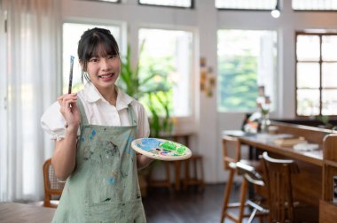 Önlüklü, elinde boya fırçası ve renk paleti olan çekici ve gülümseyen genç bir Asyalı kadın sanatçının portresi. Kendi stüdyosunda duruyor. Yaratıcılık hobisi, eğlence, sanat ilhamı