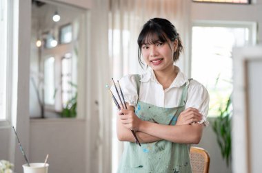 Önlüklü, güzel ve yaratıcı bir Asyalı kadın sanatçı kameraya gülümsüyor, kollarını kavuşturup duruyor ve atölyesinde elinde boya fırçaları tutuyor..