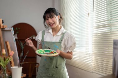 Resim stüdyosundaki tuvalde fırça ve renk paleti tutan önlüklü genç ve yaratıcı bir Asyalı kadın ressamın portresi. Sanat atölyesi, yaratıcılık hobisi