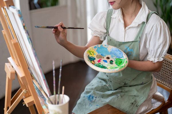 Обрезанное изображение молодой азиатской художницы, держащей цветовую палитру и кисть, рисующей на холсте акварелью в своей домашней студии. Концепция хобби творчества