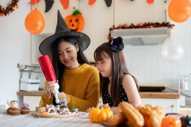 Cadı şapkalı, nazik ve güzel bir Asyalı anne, Cadılar Bayramı 'nda küçük kızıyla birlikte evde Cadılar Bayramı' nı kutlayarak kek yapmaktan zevk alıyor..