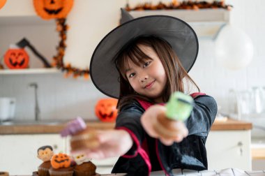 Cadılar Bayramı kostümü giymiş sevimli ve gülümseyen Asyalı bir kızın portresi, Cadılar Bayramı 'nda mutfakta kek yapmaktan zevk alıyor..