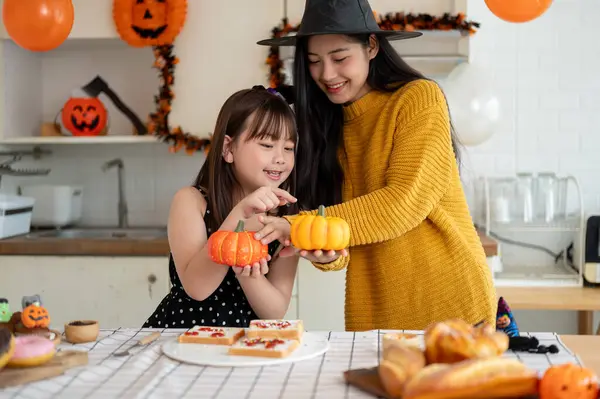 A cheerful and beautiful young Asian mom is enjoying playing with her young daughter while preparing food for Halloween in the kitchen together. Happy family, Spooky season, Special event