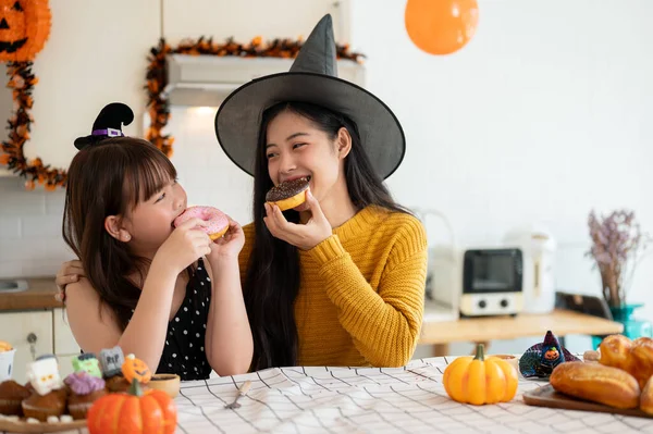 A happy and pretty young Asian girl in a Halloween costume enjoys eating doughnuts with her mom in the kitchen, having fun, celebrating Halloween with her mom at home. Happy family time, Spooky season