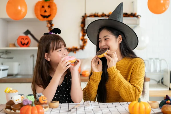 A happy and pretty young Asian girl in a Halloween costume enjoys eating doughnuts with her mom in the kitchen, celebrating Halloween with her mom at home. Happy family time, Spooky season
