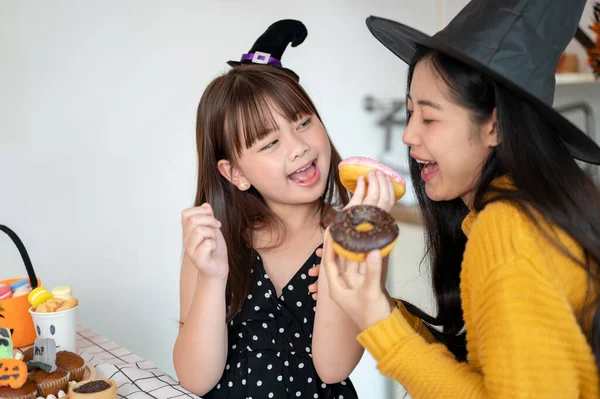 A happy and pretty young Asian girl in a Halloween costume enjoys eating doughnuts with her mom in the kitchen, having fun, celebrating Halloween with her mom at home. Happy family time, Spooky season