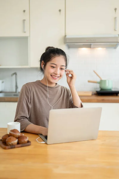 A happy and beautiful young Asian woman is working from home, wearing earphones and using her laptop at a table in the kitchen, having an online meeting. People and technology concepts