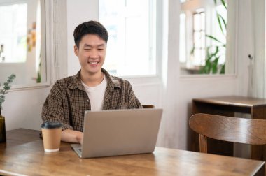 Sıradan kıyafetler giyen yakışıklı ve zeki bir Asyalı minimalist bir kahve dükkanında dizüstü bilgisayarını kullanıyor. Uzaktan çalışan, dijital göçebe, serbest çalışan, üniversite öğrencisi.