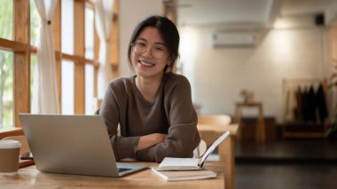 Çekici ve gülümseyen Asyalı bir kadının portresi. Gözlüklü, dizüstü bilgisayarı ve kitaplarıyla bir masada oturuyor..