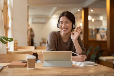 Neşeli ve çekici, kulaklık takan genç bir Asyalı kadın rahat bir kahve dükkanında dijital tabletiyle bir masada oturuyor elini sallıyor ve kameraya gülümsüyor. Uzak çalışma konsepti