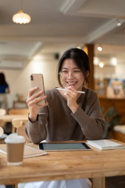 Kafedeki bir masada otururken akıllı telefonunu kullanan sevimli ve neşeli bir Asyalı kadının portresi. Video çağrısı, komik videolar izleme, sosyal medyada kayma