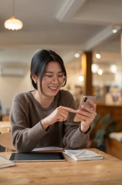 Güzel ve neşeli bir Asyalı kadın bir kahve dükkanındaki masada otururken akıllı telefonunu kullanıyor. sohbet, çevrimiçi video izleme, mesajlara cevap verme, sosyal medyada kaydırma