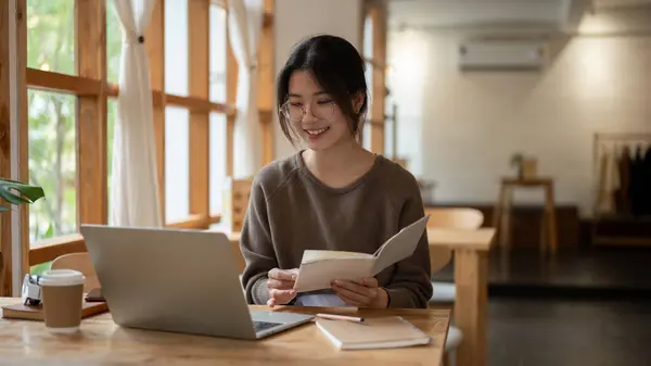 A happy and relaxed young Asian female college student working remotely at a beautiful cafe, working on her laptop, doing homework.