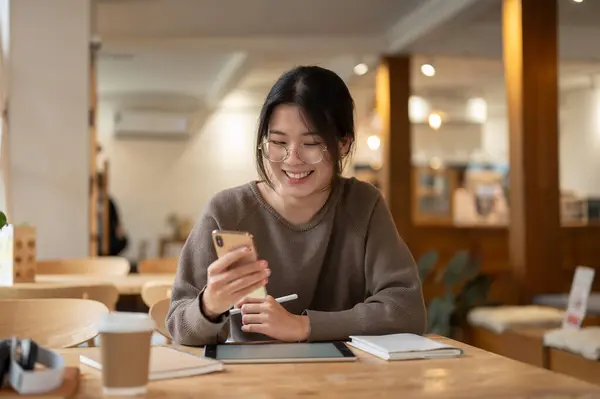 A cheerful and pretty Asian woman enjoys chatting with her friends through her smartphone while sitting at a table in a coffee shop.