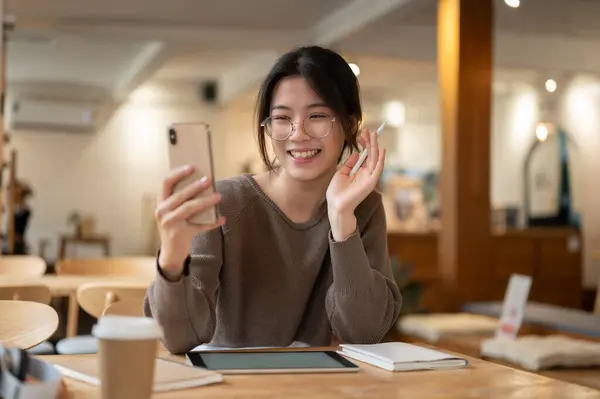 A cheerful and pretty Asian woman enjoys talking on a video call with her friends through her smartphone while sitting at a table in a coffee shop. Lifestyle concept
