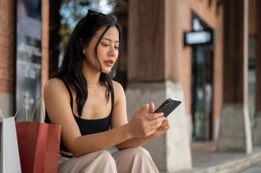 Harika bir Asyalı kadın telefonunu kullanıyor, arkadaşlarıyla sohbet ediyor. Alışveriş torbalarıyla perakende mağazasının önünde dinleniyor..