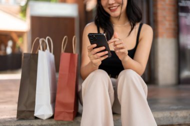 Harika bir Asyalı kadın telefonunu kullanıyor, alışveriş torbalarıyla perakende satış mağazasının merdivenlerinde dinlenirken arkadaşlarıyla sohbet ediyor. kırpılmış resim