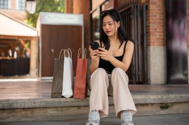 Harika bir Asyalı kadın telefonunu kullanıyor, alışveriş torbalarıyla perakende satış mağazasının merdivenlerinde dinlenirken arkadaşlarıyla sohbet ediyor. Yaşam tarzı ve alışveriş çılgınlığı kavramları
