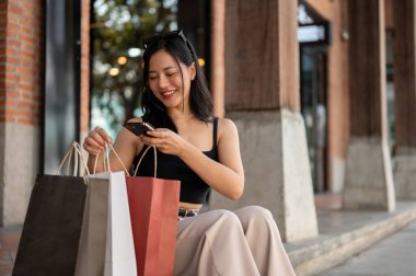 Muhteşem bir Asyalı kadın perakende mağazasının önündeki merdivenlerde dinlenirken alışveriş torbalarının fotoğraflarını çekiyor. Yaşam tarzı ve alışveriş çılgınlığı kavramları
